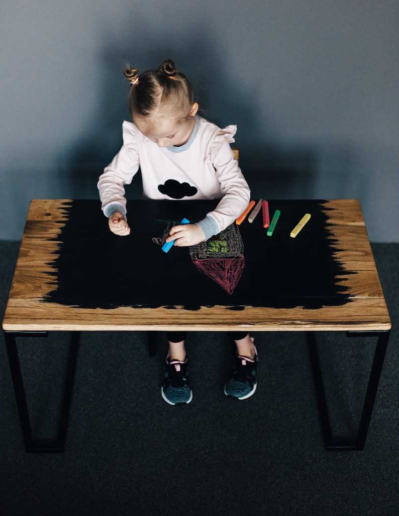 May include: A young child sits at a black chalkboard table drawing with colorful chalk. The table has a wooden top and black metal legs.