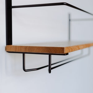 K&ouml;nnte beinhalten: Ein Wandregal aus Holz mit einem schwarzen Metallrahmen und zwei Haken darunter. Das Regal ist aus hellem Holz und der Rahmen aus schwarzem Metall.