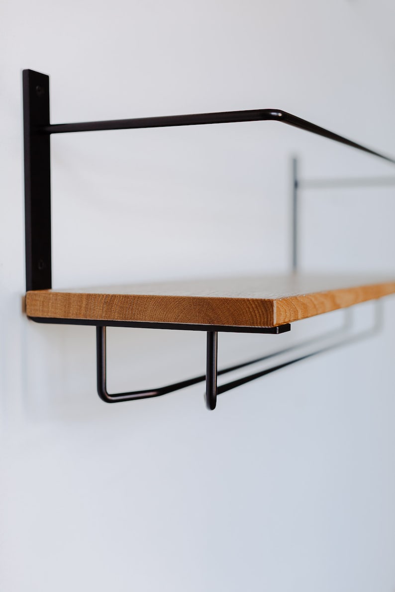 K&ouml;nnte beinhalten: Ein Wandregal aus Holz mit einem schwarzen Metallrahmen und zwei Haken darunter. Das Regal ist aus hellem Holz und der Rahmen aus schwarzem Metall.