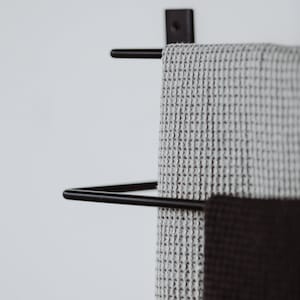 Pode incluir: Um suporte de toalha de metal preto com duas barras, segurando uma toalha de waffle cinza e cinza escuro.
