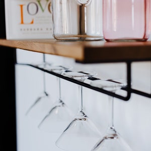 May include: A set of five clear glass martini glasses hanging upside down from a black metal rack. The rack is mounted on a wooden shelf.