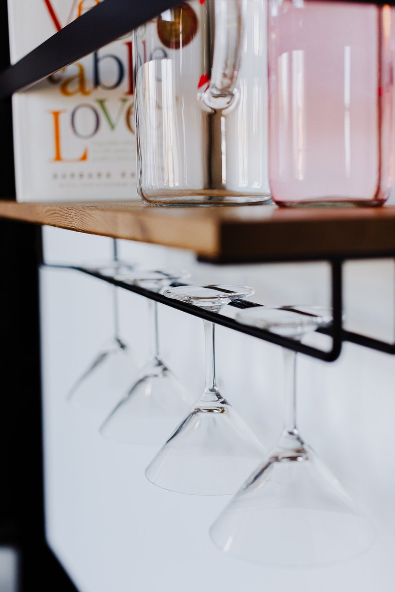 May include: A set of five clear glass martini glasses hanging upside down from a black metal rack. The rack is mounted on a wooden shelf.