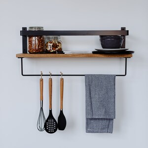 May include: A black metal shelf with a wooden top and a towel bar. The shelf has two glass jars filled with nuts and a plate with bowls on top. There are three wooden handled kitchen utensils hanging from the towel bar.