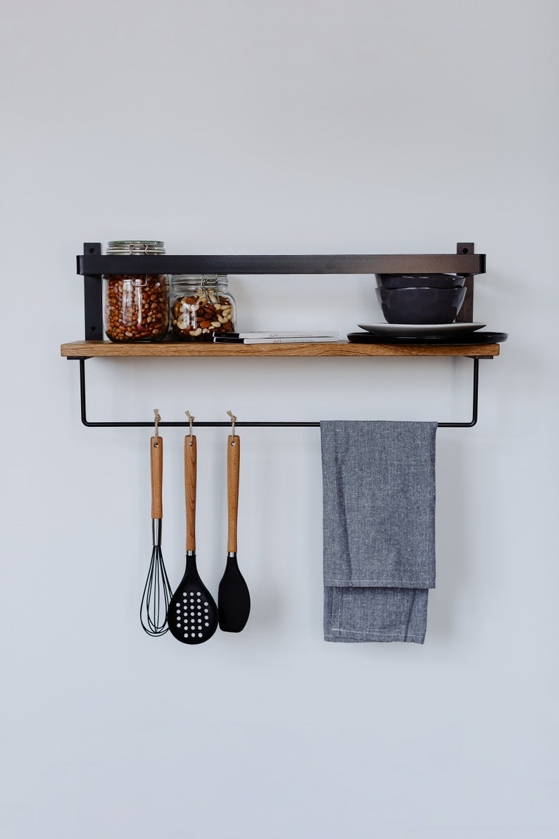 May include: A black metal shelf with a wooden top and a towel bar. The shelf has two glass jars filled with nuts and a plate with bowls on top. There are three wooden handled kitchen utensils hanging from the towel bar.