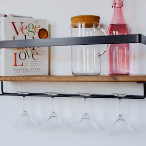 May include: A black metal shelf with a wooden top, featuring a glass pitcher with a cork lid, a pink glass bottle, and four hanging wine glasses. The shelf also holds a stack of books, including one titled "Veggie Love" by Barbara Kafka.