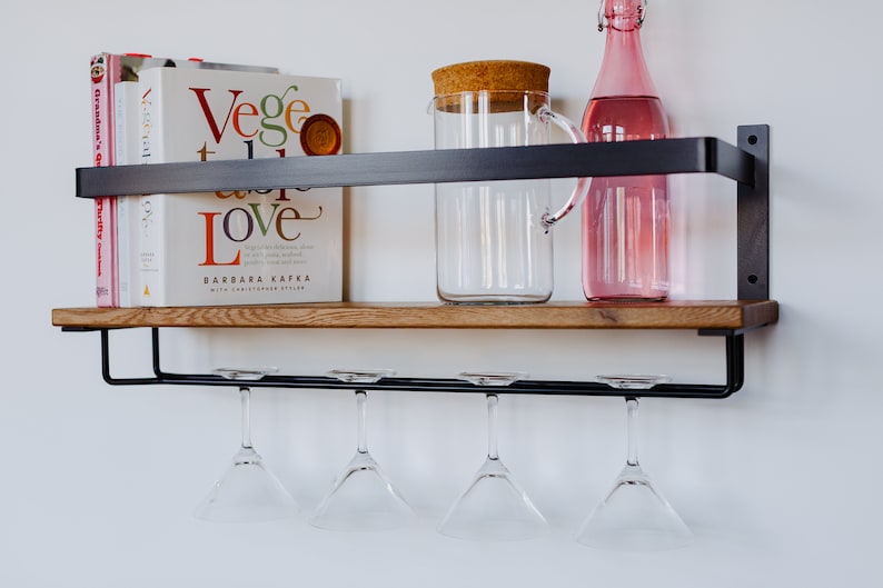 May include: A black metal shelf with a wooden top, featuring a glass pitcher with a cork lid, a pink glass bottle, and four hanging wine glasses. The shelf also holds a stack of books, including one titled "Veggie Love" by Barbara Kafka.