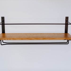 K&ouml;nnte beinhalten: Ein Holzregal mit einem schwarzen Metallrahmen und zwei schwarzen Metallstangen darunter. Das Regal ist an einer wei&szlig;en Wand montiert.