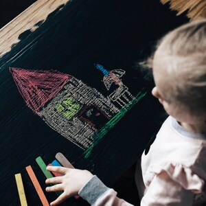 May include: A child draws a house with a red roof and a green lawn on a black chalkboard.  The child is holding a set of colorful chalk sticks.