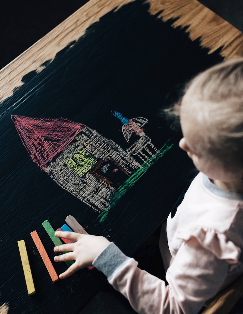 May include: A child draws a house with a red roof and a green lawn on a black chalkboard.  The child is holding a set of colorful chalk sticks.