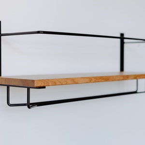 K&ouml;nnte beinhalten: Ein schwarzes Wandregal aus Metall mit einer Holz-Oberplatte und einer Metallstange darunter. Das Regal ist an einer wei&szlig;en Wand montiert.