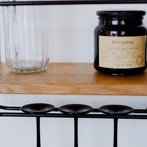 K&ouml;nnte beinhalten: Ein schwarzer Metall-Weinregal mit drei Glashaltern und einem Holzregal. Eine klare Glasflasche und ein braunes Glasgef&auml;&szlig; mit einem Etikett mit der Aufschrift "Mahogany Scented Wax Candle" stehen auf dem Regal.