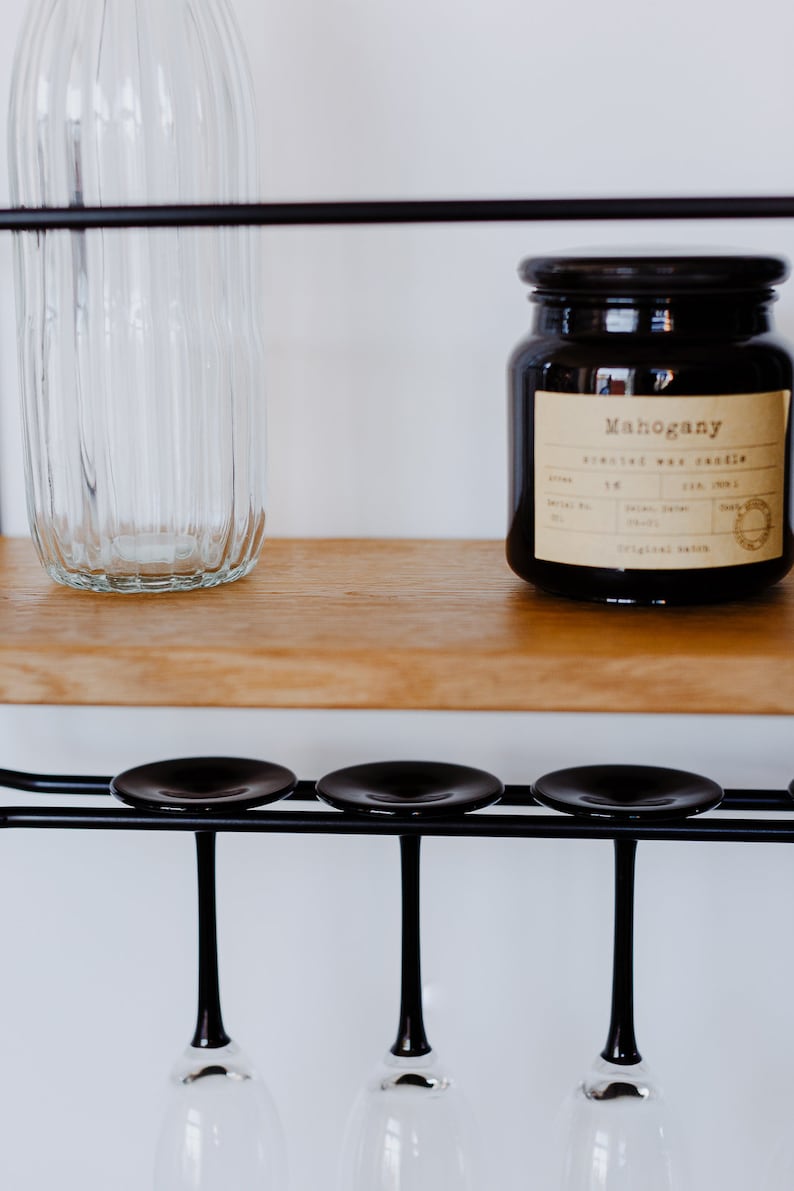 K&ouml;nnte beinhalten: Ein schwarzer Metall-Weinregal mit drei Glashaltern und einem Holzregal. Eine klare Glasflasche und ein braunes Glasgef&auml;&szlig; mit einem Etikett mit der Aufschrift "Mahogany Scented Wax Candle" stehen auf dem Regal.