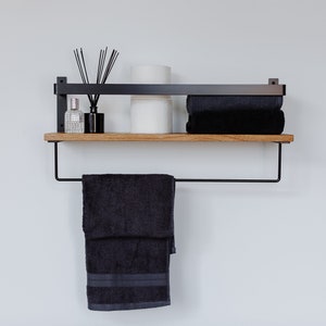 May include: A black metal shelf with a wooden top, featuring a towel bar and two shelves. The top shelf holds a stack of towels, a roll of toilet paper, and a bottle of diffuser oil with reeds. The bottom shelf is empty.