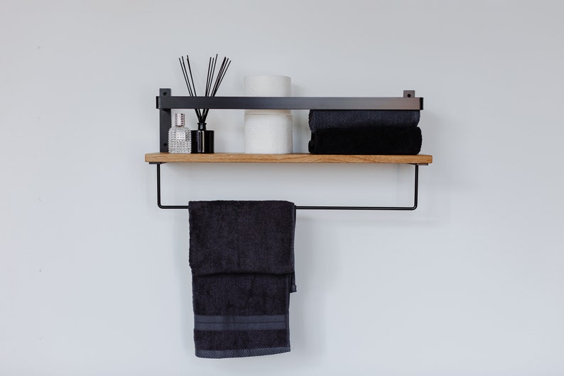 May include: A black metal shelf with a wooden top, featuring a towel bar and two shelves. The top shelf holds a stack of towels, a roll of toilet paper, and a bottle of diffuser oil with reeds. The bottom shelf is empty.