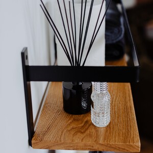 May include: A black metal shelf with a wooden top. The shelf has a black metal rod hanging down from the bottom. There are two bottles on the shelf, one black and one clear. A diffuser with black sticks is also on the shelf.