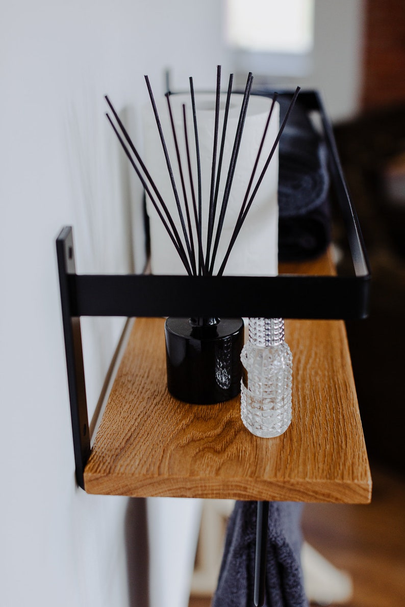 May include: A black metal shelf with a wooden top. The shelf has a black metal rod hanging down from the bottom. There are two bottles on the shelf, one black and one clear. A diffuser with black sticks is also on the shelf.