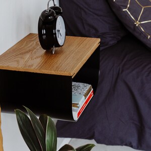 May include: A black metal floating nightstand with a wood top. The nightstand has a shelf underneath for storage. A black alarm clock is on the top shelf and a stack of books is on the bottom shelf.
