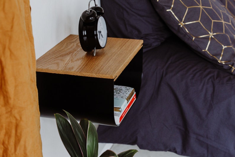 May include: A black metal floating nightstand with a wood top. The nightstand has a shelf underneath for storage. A black alarm clock is on the top shelf and a stack of books is on the bottom shelf.