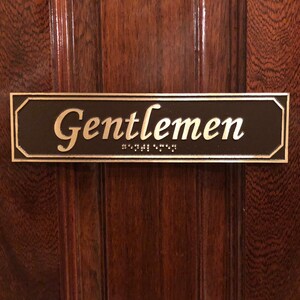 May include: A brown rectangular sign with rounded corners and a silver border. The sign reads "Gentlemen" in a gold cursive font with braille lettering below.