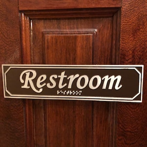 May include: A brown rectangular sign with silver lettering that reads "Restroom" with braille underneath.