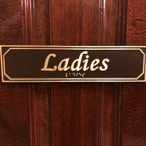 May include: A brown rectangular sign with a gold border and the word "Ladies" in gold lettering. The sign also has braille lettering.