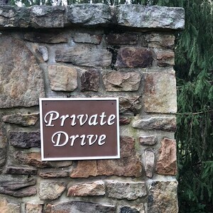 以下が含まれることがあります： 「Private Drive」と書かれた茶色の看板がついた石の柱。柱は積み重ねられた石でできており、緑の木々と木製のフェンスに囲まれている。