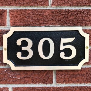 May include: A black and gold house number plaque with the number 305 in white lettering. The plaque is mounted on a brick wall.