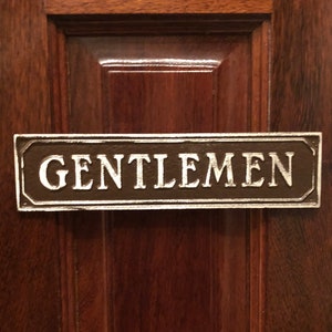 May include: A brown wooden door with a metal sign that reads "GENTLEMEN" in white lettering.