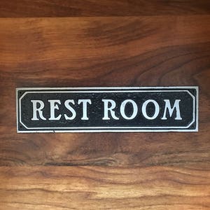 Pu&ograve; includere: Un cartello nero e argento con la scritta "REST ROOM" in lettere bianche.