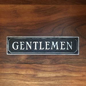 May include: A black metal sign with silver lettering that reads "GENTLEMEN".