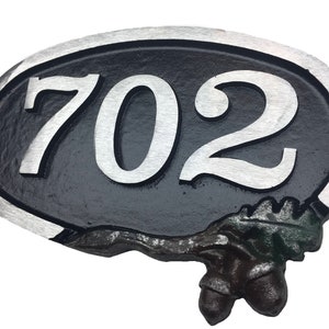 May include: A black oval-shaped house number sign with a silver border and silver numbers "702". The sign has a decorative acorn and leaf design at the bottom.