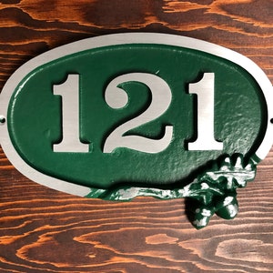 May include: Oval-shaped house number sign with a green background and silver numbers. The numbers "121" are displayed in a bold, sans-serif font. The sign has a silver border and a decorative acorn and leaf design at the bottom.