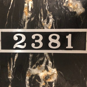 May include: Rectangular black address plaque with the numbers "2381" in a silver font. The plaque has a silver border and is mounted on a black and gold marble-like surface. Two screws secure the plaque.