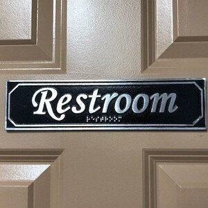 May include: A black and silver sign with the word "Restroom" in silver lettering on a beige door.