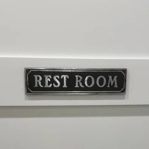 Pu&ograve; includere: Una porta bianca con una maniglia nera e un cartello nero con la scritta "REST ROOM" in lettere argentate.