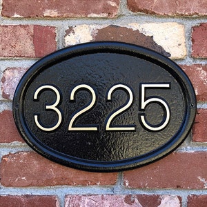 May include: Black oval-shaped house number sign with the number 3225 in gold lettering on a brick wall.