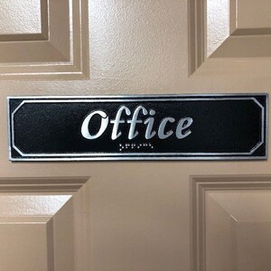 May include: A black and silver rectangular sign with the word "Office" in silver lettering. The sign has braille lettering below the word "Office".