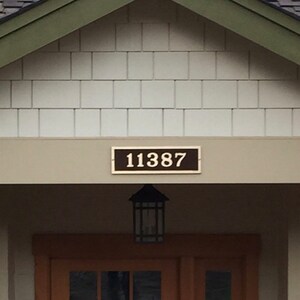 May include: A white house with a brown roof and a wooden door. The house has a green trim around the roof and a white porch with two columns. The house number is 11387.
