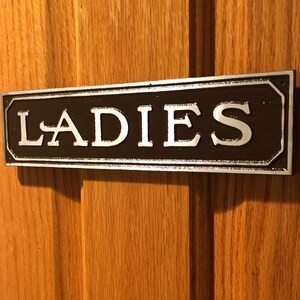 K&ouml;nnte beinhalten: Ein schwarzes und silbernes Schild mit dem Wort "LADIES" in wei&szlig;en Buchstaben.