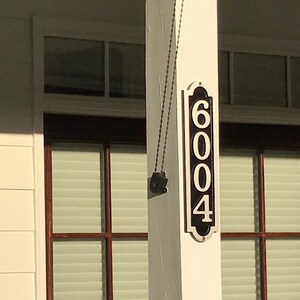 May include: A white house with a black porch and a black hanging house number sign that reads "6004". There is a black hanging light fixture on the side of the house and a hanging planter with red flowers.
