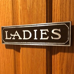 May include: A black and silver metal sign with the word "LADIES" in white lettering.