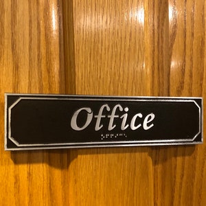 May include: A black and silver rectangular sign with the word "Office" in silver lettering. The sign also includes braille lettering.