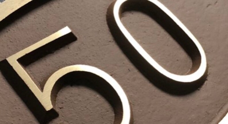 May include: Close-up of a gold-colored metal number "50" on a brown background.