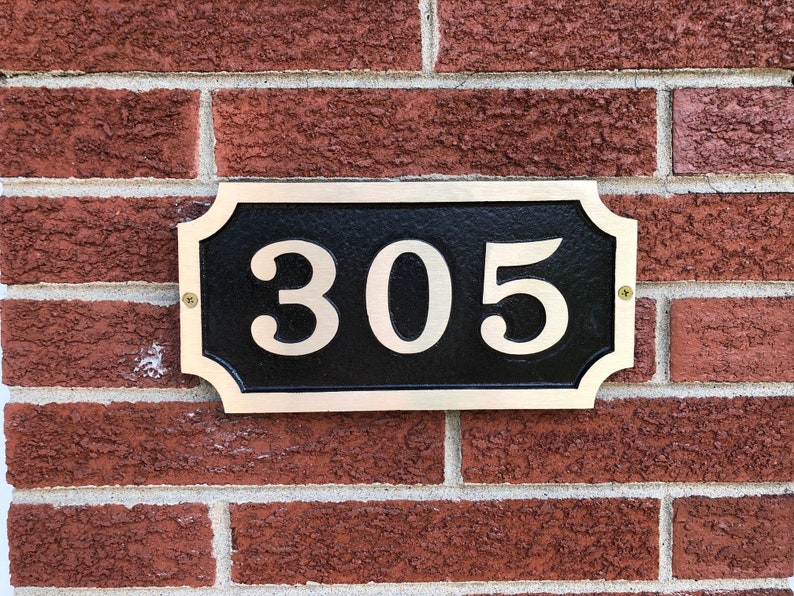 May include: A black and gold house number plaque with the number 305 in white lettering. The plaque is mounted on a brick wall.