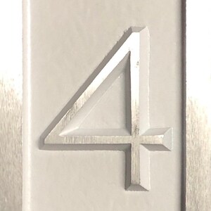 May include: A close-up of a white, rectangular sign with the number "4" embossed in the center. The number is raised and has a slightly metallic sheen.