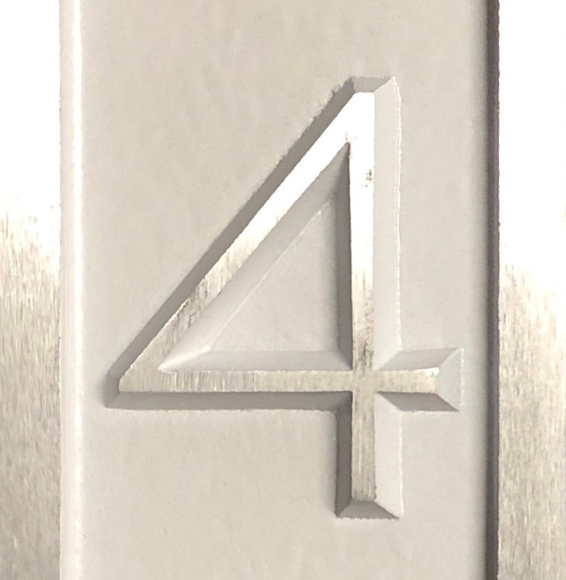 May include: A close-up of a white, rectangular sign with the number "4" embossed in the center. The number is raised and has a slightly metallic sheen.