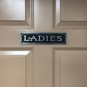 May include: A beige door with a black and silver sign that reads "LADIES".