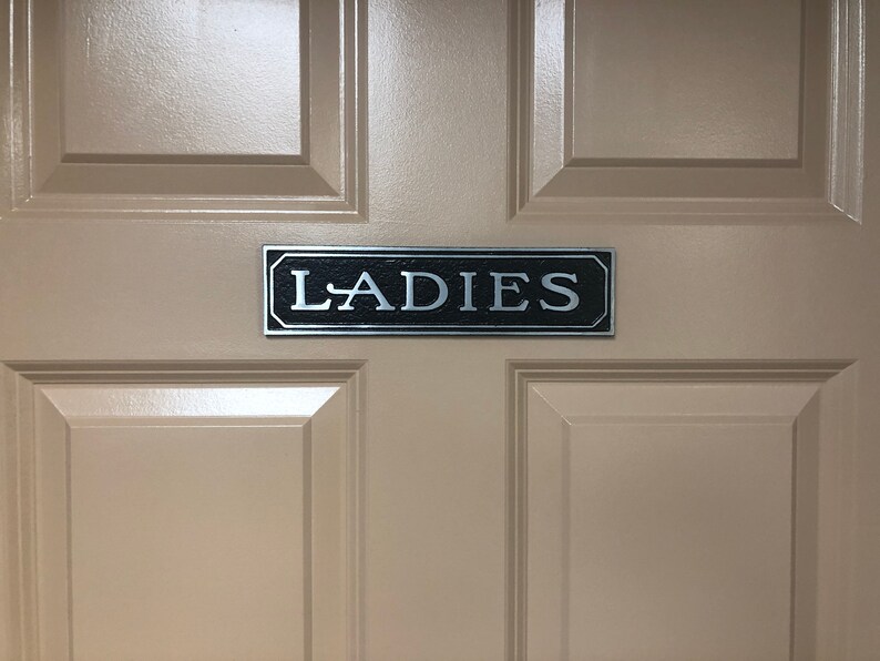 May include: A beige door with a black and silver sign that reads "LADIES".