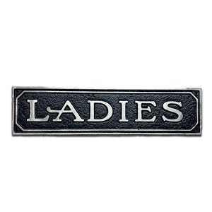 K&ouml;nnte beinhalten: Ein schwarzes und silbernes Metallschild mit dem Wort "LADIES" in wei&szlig;en Buchstaben.