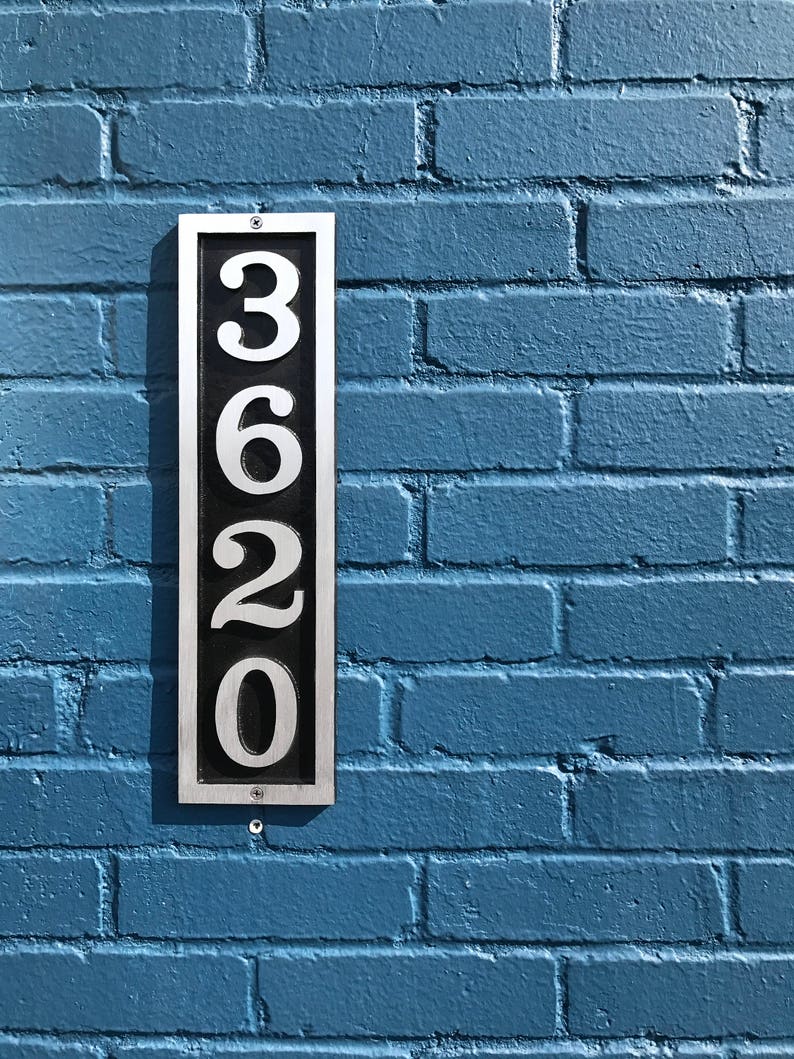 Puede incluir: Una placa de n&uacute;mero de casa de metal plateado con fondo negro y n&uacute;meros blancos que indican 3620, montada en una pared de ladrillo azul.